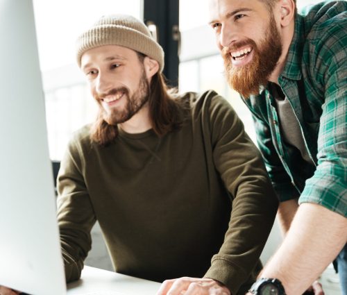 Photo of young happy men colleagues in office using computer. Looking aside.